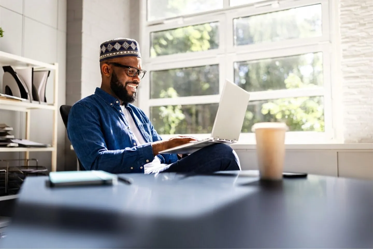 Smiling Muslim man in a kufi and glasses working on a laptop at a desk by a bright window with coffee beside him.